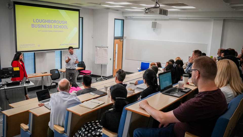 People seated in a tiered classroom, facing a lecturer with a projector screen displaying 